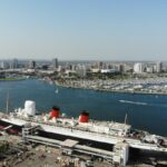 Vista aérea do navio Queen Mary atracado no porto de Long Beach, com o horizonte da cidade e vários barcos na marina ao fundo.