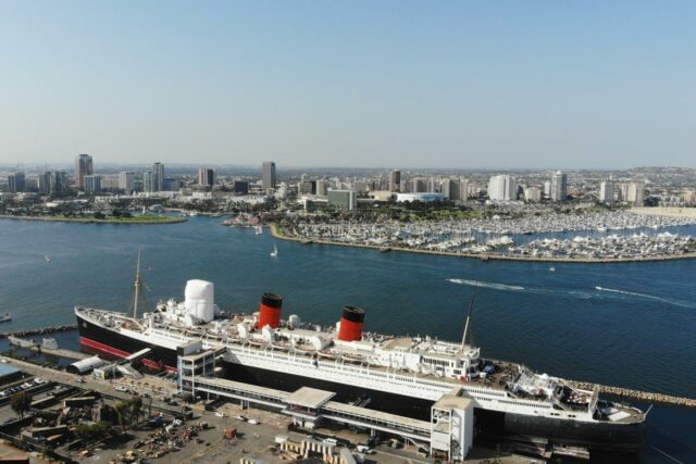 Vista aérea do navio Queen Mary atracado no porto de Long Beach, com o horizonte da cidade e vários barcos na marina ao fundo.