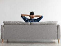 Casal chocado com o que encontraram escondido em seu sofá novo Vista traseira de um homem de camisa xadrez relaxando em um sofá cinza com as mãos atrás da cabeça.