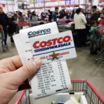 Uma mão segurando um cartão Costco Gold Star Member em frente a uma placa borrada da loja Costco Wholesale.