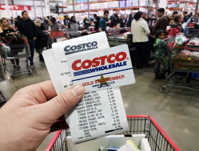 Uma mão segurando um cartão Costco Gold Star Member em frente a uma placa borrada da loja Costco Wholesale.