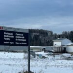 Um homem na faixa dos 30 ou 40 anos, com barba clara, usando boné e capa de chuva cinza, está em frente a um rio calmo com grama cor de palha ao longo das margens e um céu cinza ao fundo.