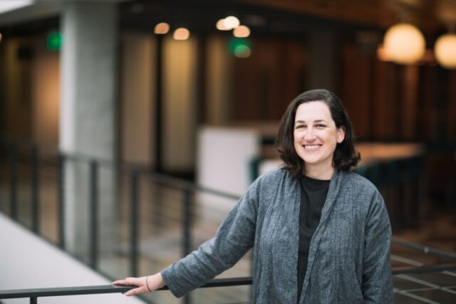 A dark-haired woman poses by a balcony in a gray cardigan sweater.