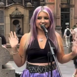 Uma jovem de cabelo roxo canta em um microfone enquanto toca na George Street, em Sydney. Ela está vestindo uma blusa preta e saia roxa.