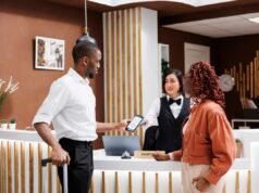 Cuidado com essas 5 cobranças ocultas de hotéis – enquanto o prefeito de Nova York, Zohran Mamdani, impõe a proibição de ‘taxas indesejadas’ sorrateiras Um turista usa seu celular para pagar na recepção de um hotel, enquanto uma recepcionista asiática segura um terminal de pagamento.