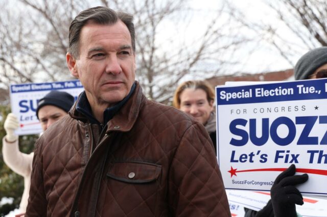 Tom Suozzi vestindo uma jaqueta acolchoada marrom, com uma placa de campanha Suozzi For Congress ao fundo.