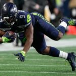 Um jogador do Seattle Seahawks com uniforme azul escuro correndo com a bola de futebol durante um jogo.