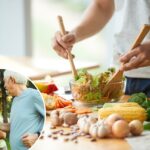 Homem misturando uma salada de legumes com utensílios de madeira em uma tigela de vidro.
