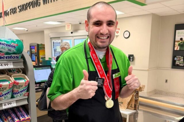 Michael Masterangelo no Publix, vestindo camisa verde, avental preto e medalha da Special Olympics, fazendo sinal de positivo.
