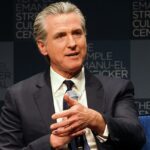 A água flui de cinco portões na represa Shasta, com um lago atrás e montanhas nevadas ao longe.