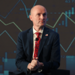 Governador de Utah intensifica briga sobre regulamentação do mercado de previsão com o presidente da CFTC. Um homem careca, de terno azul-marinho e gravata estampada vermelha, está sentado em um palco com um microfone preso à jaqueta, com as mãos entrelaçadas, em frente a um cenário que exibe gráficos financeiros e gráficos de velas.