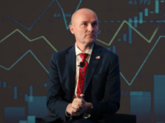 Governador de Utah intensifica briga sobre regulamentação do mercado de previsão com presidente da CFTC Governador de Utah intensifica briga sobre regulamentação do mercado de previsão com o presidente da CFTC. Um homem careca, de terno azul-marinho e gravata estampada vermelha, está sentado em um palco com um microfone preso à jaqueta, com as mãos entrelaçadas, em frente a um cenário que exibe gráficos financeiros e gráficos de velas.