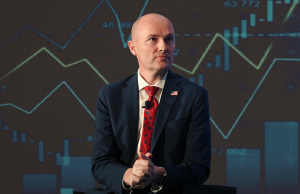 Governador de Utah intensifica briga sobre regulamentação do mercado de previsão com presidente da CFTC Governador de Utah intensifica briga sobre regulamentação do mercado de previsão com o presidente da CFTC. Um homem careca, de terno azul-marinho e gravata estampada vermelha, está sentado em um palco com um microfone preso à jaqueta, com as mãos entrelaçadas, em frente a um cenário que exibe gráficos financeiros e gráficos de velas.