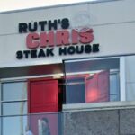 Ruth's Chris Steak House com entrada dupla vermelha e duas pessoas em pé em uma varanda abaixo do restaurante.