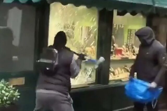 Dois ladrões mascarados assaltando uma joalheria, um quebrando a vitrine com uma ferramenta, o outro segurando uma bolsa azul.