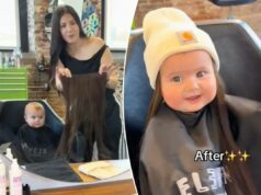 Mamãe dá longas extensões de cabelo preto à adorável filha de 8 meses – veja como o bebê reage Uma mulher segurando extensões de cabelo em frente a um espelho, com um bebê com uma capa de barbeiro refletido no espelho.