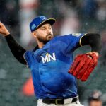 Um jogador de beisebol com boné preto dos Twins e camisa preta com uma luva vermelha chega ao campo.
