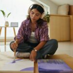 Uma jovem com fones de ouvido sentada no chão, pintando em uma tela com tinta roxa.