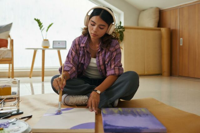 Uma jovem com fones de ouvido sentada no chão, pintando em uma tela com tinta roxa.
