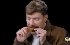 O episódio de MrBeasts Hot Ones é doloroso de assistir Um homem está sentado em um quarto escuro mordendo uma asa de frango.