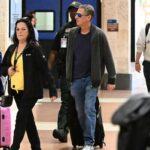 Michael Feldman, marido de Savannah Guthrie, caminhando por um aeroporto da Flórida com uma mala rosa, seguido por uma criança com fones de ouvido lilás e um policial.