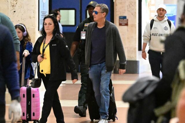 Michael Feldman, marido de Savannah Guthrie, caminhando por um aeroporto da Flórida com uma mala rosa, seguido por uma criança com fones de ouvido lilás e um policial.