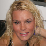 Kirsty Wilkinson usa um vestido prateado de manga curta com um padrão de flores pretas. Ela é branca, com cabelos loiros descoloridos que chegam abaixo dos ombros. Ela está com os braços levantados e sorrindo para a câmera. Ela usa uma pulseira larga de prata. Ao fundo, que é à noite, uma placa diz "Comida Fish'n'Chips para levar" está iluminado. Há um poste amarelo atrás dela e a borda de um dossel. Parece um local de feira.