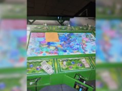 Oito presos enquanto cassino ilegal é fechado pela polícia na Pensilvânia Oito presos porque um cassino ilegal foi fechado pela polícia na Pensilvânia. Close de uma mesa de jogo de cassino ilegal apreendida durante uma operação policial, mostrando uma superfície de jogo colorida com gráficos com tema subaquático, dinheiro espalhado, fichas estilo pôquer e pequenas estatuetas, com partes da máquina abertas e fiação exposta, indicando que ela foi desligada e confiscada.