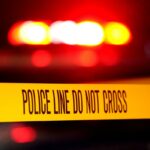 A linha policial não cruza a fita em uma cena de crime com luzes vermelhas piscando ao fundo.