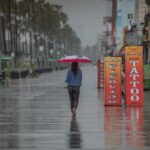 Um homem vestindo um poncho azul e um boné de beisebol anda de skate em um calçadão molhado, com palmeiras e edifícios ao fundo durante fortes chuvas.