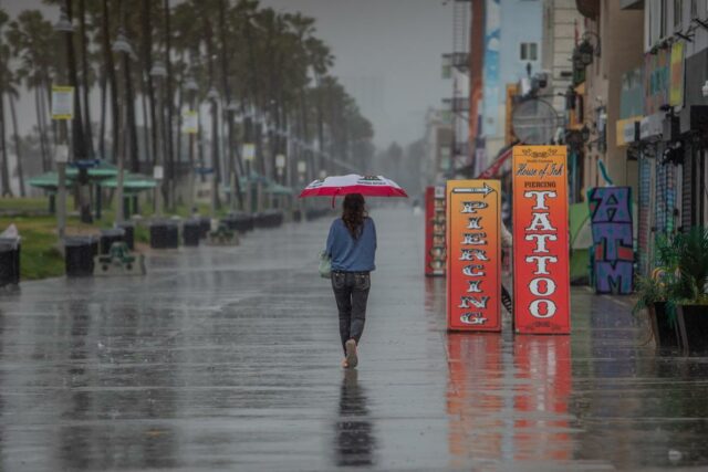 Um homem vestindo um poncho azul e um boné de beisebol anda de skate em um calçadão molhado, com palmeiras e edifícios ao fundo durante fortes chuvas.