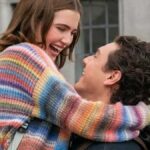 Uma mulher loira está rindo e segurando um cachorrinho vestido com uma roupa roxa e rosa. Ela está vestida com uma jaqueta rosa brilhante, óculos escuros vermelhos e um telefone rosa preso à orelha. O fundo mostra um ambiente externo desfocado com pessoas e vegetação.