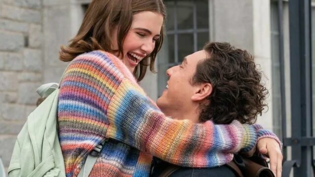 Uma mulher loira está rindo e segurando um cachorrinho vestido com uma roupa roxa e rosa. Ela está vestida com uma jaqueta rosa brilhante, óculos escuros vermelhos e um telefone rosa preso à orelha. O fundo mostra um ambiente externo desfocado com pessoas e vegetação.