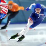 Jordan Stolz, da equipe de patinação de velocidade dos Estados Unidos, vestindo um terno azul, branco e vermelho com outro patinador de velocidade desfocado ao fundo durante a prova masculina de 500m.