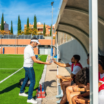 O regulador francês de jogos de azar, ANJ, alerta sobre preocupações crescentes com anúncios de apostas na Copa do Mundo. Um treinador de futebol distribui garrafas de água para jogadores que usam babadores vermelhos enquanto eles se sentam no banco durante um intervalo em um campo ensolarado ao ar livre, com um jogador próximo segurando garrafas adicionais.