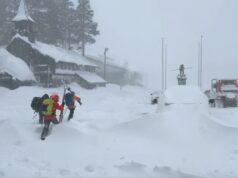 Seis esquiadores resgatados após serem enterrados em uma avalanche devastadora perto do Lago Tahoe: policiais Dois indivíduos com roupas de inverno caminhando na neve profunda, passando por um prédio coberto de neve, um gato da neve e carros enterrados.