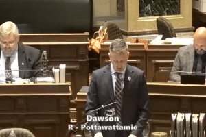 O Senado de Iowa avança com medidas abrangentes de aplicação de jogos de azar e mudanças fiscais em um projeto de lei importante. Um homem de terno está em um pódio no plenário do Senado de Iowa, lendo comentários preparados, com outros dois homens de terno sentados atrás dele em suas mesas; o texto na tela o identifica como “Dawson R - Pottawattamie”.