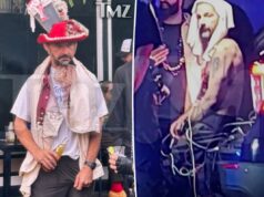 Shia LaBeouf retorna imediatamente às festas do Mardi Gras após ser libertado da prisão Shia LaBeouf, parecendo embriagado, vestido com um chapéu vermelho e branco com livro falso e acabamento fofo, camiseta branca, colete vermelho e bege e calça escura, segurando uma garrafa de cerveja.