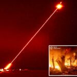 Um raio laser vermelho brilhante sai de um lançador terrestre em direção a um alvo no céu noturno.