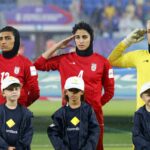 Três jogadoras de futebol iranianas fazem continência com três crianças na frente delas, enquanto chove.