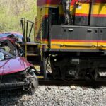 Uma van vermelha gravemente danificada ao lado de um trem de carga nos trilhos.