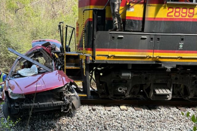 5 mortos após trem de carga atingir van em cruzamento Uma van vermelha gravemente danificada ao lado de um trem de carga nos trilhos.