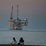 Plataforma de petróleo offshore Esther em Seal Beach, Califórnia, com duas pessoas sentadas na praia em primeiro plano e gaivotas voando.