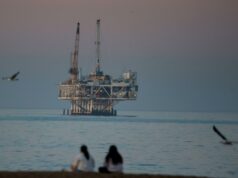 A Califórnia precisa de petróleo. Deixe fluir – tão constante quanto o bom senso Plataforma de petróleo offshore Esther em Seal Beach, Califórnia, com duas pessoas sentadas na praia em primeiro plano e gaivotas voando.