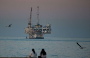 A Califórnia precisa de petróleo. Deixe fluir – tão constante quanto o bom senso Plataforma de petróleo offshore Esther em Seal Beach, Califórnia, com duas pessoas sentadas na praia em primeiro plano e gaivotas voando.