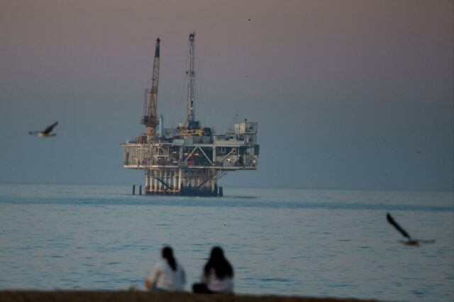 Plataforma de petróleo offshore Esther em Seal Beach, Califórnia, com duas pessoas sentadas na praia em primeiro plano e gaivotas voando.