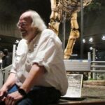 Uma mulher sorridente e tatuada com cabelo roxo claro, boné de beisebol e piercing no septo tira uma selfie ao ar livre ao lado de formações rochosas no deserto