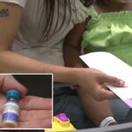 Uma pessoa com manga preta tocando a testa de uma criança deitada em uma cama de hospital.