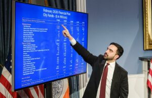 A empresa de classificação de crédito S&P torna-se a segunda a soar o alarme sobre as finanças de Nova York sob Mamdani Homem de terno apontando para uma tela grande exibindo o Plano Financeiro de fevereiro de 2026 para os Fundos da Cidade de Nova York.