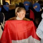 Bonnie Blue (Tia Billinger) sorrindo em frente à Embaixada da Indonésia no Reino Unido, cercada por pessoas com balaclavas azuis e segurando a bandeira da Indonésia.